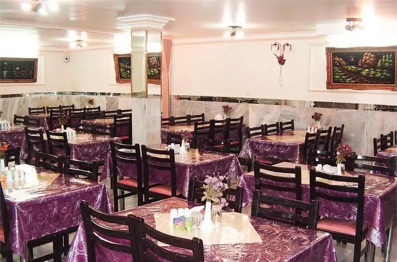 نمای داخلی رستوران ایرانی پذیرا با میز و صندلی‌های چوبی، رومیزی‌های بنفش و دکوراسیون سنتی و شیک برای پذیرایی از مشتریان.