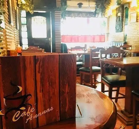 محیط دلنشین و گرم کافه آناناس با دکوراسیون چوبی، میز و صندلی‌های راحت، و منوی رومیزی با لوگوی "Cafe Ananas".