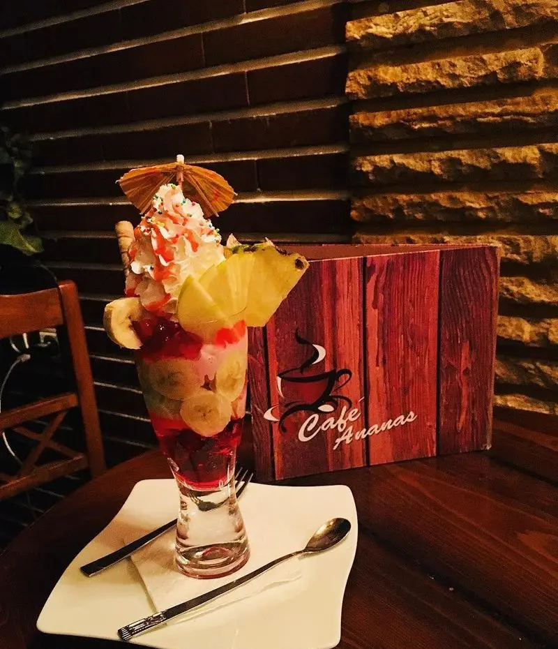 بستنی میوه ای خاص کافه آناناس با موز، آناناس، خامه و تزیینات جذاب، سرو شده روی میز چوبی در فضایی دلنشین و دنج.