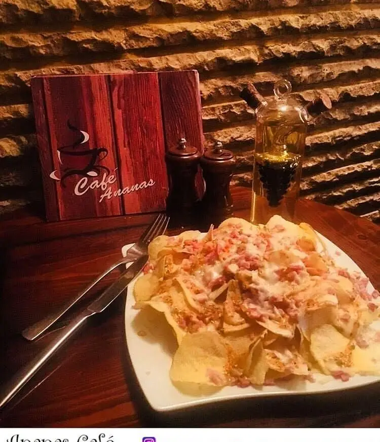 میز غذای کافه آناناس: بشقاب بزرگ چیپس سیب زمینی لود شده با سس سفید و بیکن، در کنار منو، فلفل‌دان و بطری روغن زیتون. انتخابی عالی برای اسنک یا شام.