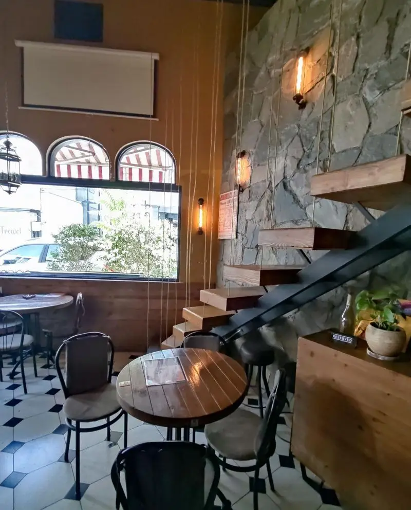 تصویری از فضای دلنشین کافه کباب آرمنیا: دیوار سنگی، پله‌های چوبی شناور، میز و صندلی‌های چوبی، کفپوش طرحدار و منظره خیابان از پنجره.