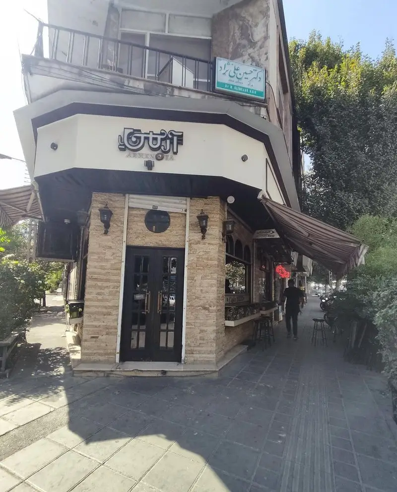 نمای خارجی کافه کباب آرمنیا، تابلوی روشن Armenia، درب ورودی مشکی و نمای سنگی. پیاده‌روی شلوغ با سایه‌انداز و میزهای فضای باز.