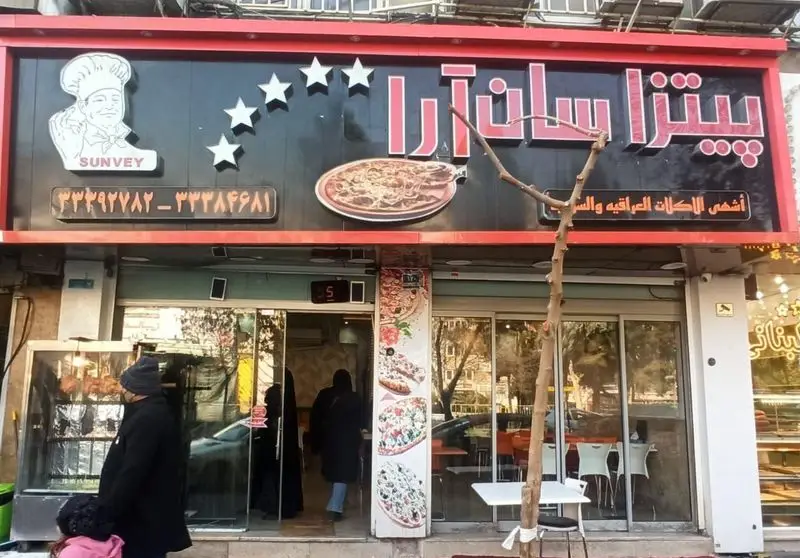 نمای بیرونی و تابلو بزرگ فست فود سان وی (پیتزا سان آرا) با لوگوی سرآشپز، ستاره و پیتزا. ارائه دهنده شاورما و غذاهای اصیل عراقی و سوری.