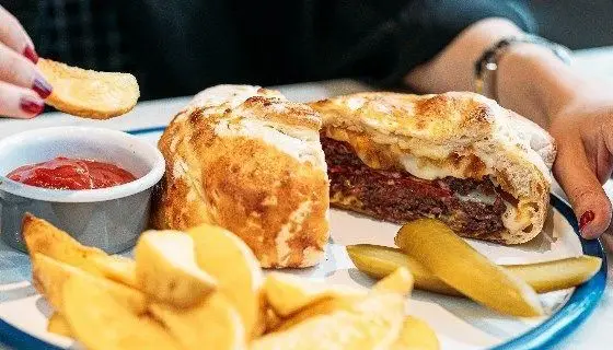 یک برگر ویژه و خوشمزه رستوران دست، نصف شده و مملو از پنیر ذوب شده و مواد تازه. در کنار سیب زمینی وج، سس کچاپ و خیارشور سرو شده است.