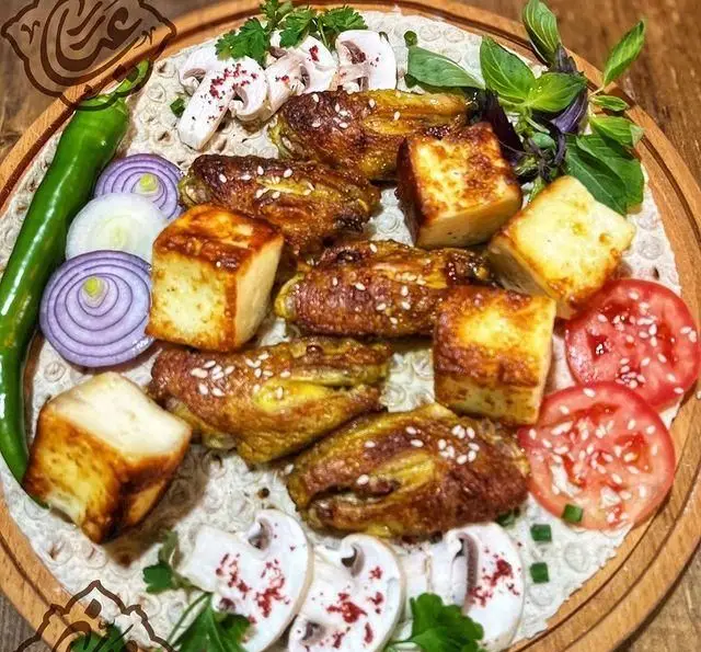 سینی جذاب کباب بال مرغ و پنیر کبابی مخصوص جگر سرای عیاران، همراه با دورچین سبزیجات تازه، قارچ، گوجه، پیاز و فلفل سبز روی نان لواش.