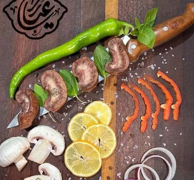 آماده‌سازی مواد اولیه تازه جگر سرای عیاران: جگر، فلفل، قارچ، لیمو، پیاز و سبزیجات معطر روی تخته آشپزی چوبی.