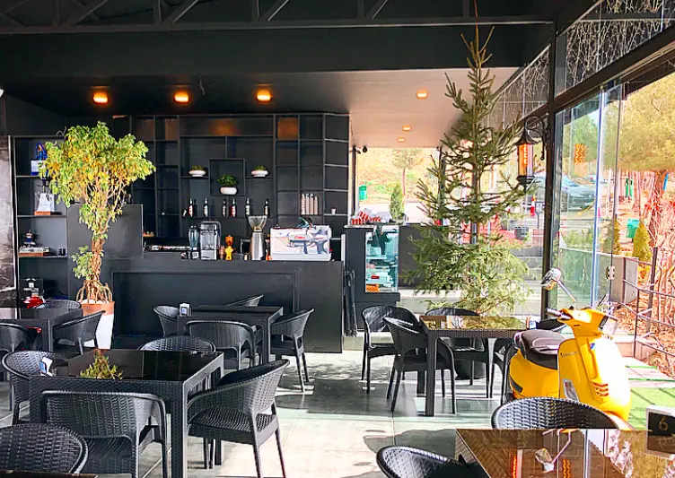 کافه رستوران لمیاء: فضای داخلی مدرن و دلنشین با میز و صندلی‌های شیک، بار مجهز، گیاهان زینتی، درخت کاج و اسکوتر وسپا زرد.