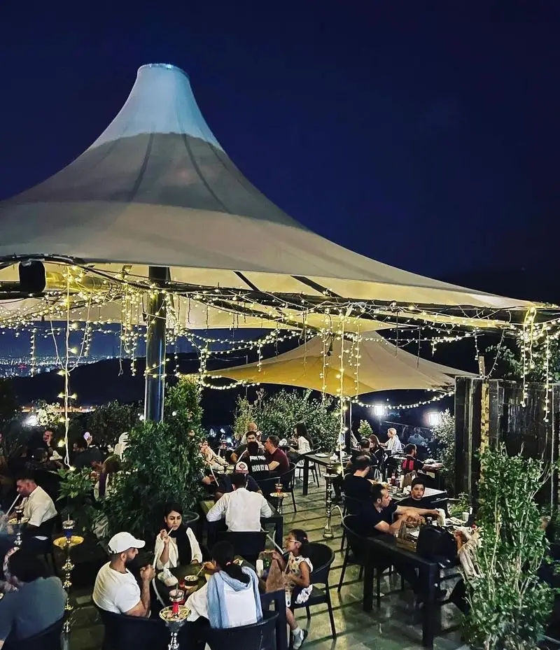 فضای شبانه کافه رستوران لمیاء با مشتریان در حال لذت بردن از غذا و قلیان، زیر نور ریسه‌های جذاب و منظره زیبای شهر.