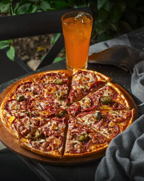 پیتزا پپرونی و هالوپینو با پنیر آب شده، برش خورده روی تخته چوبی در کافه رستوران ریون همراه نوشیدنی خنک پرتقالی.