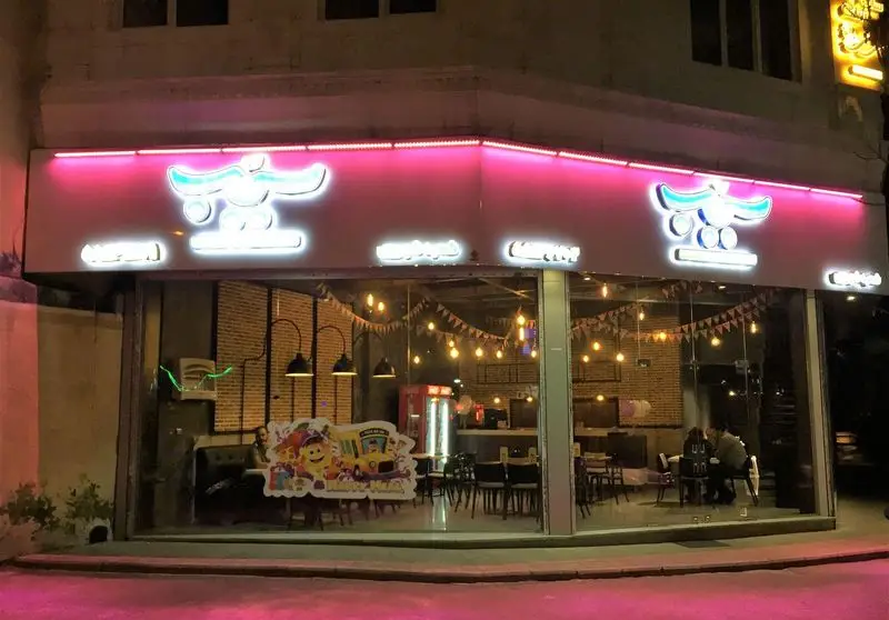 نمای بیرونی روشن و دیدنی فست فود پیتزا سیب ۳۶۰ در شب، با تابلوهای نئونی صورتی درخشان و دکوراسیون داخلی مدرن و صمیمی مناسب خانواده‌ها و مشتریان.