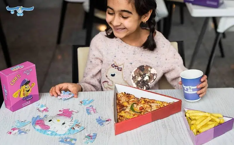 دختر شاد با پیتزا و سیب‌زمینی سرخ‌کرده پیتزا سیب ۳۶۰ در جعبه‌های مثلثی، در حال بازی با پازل Hello Kitty و نوشیدن است.