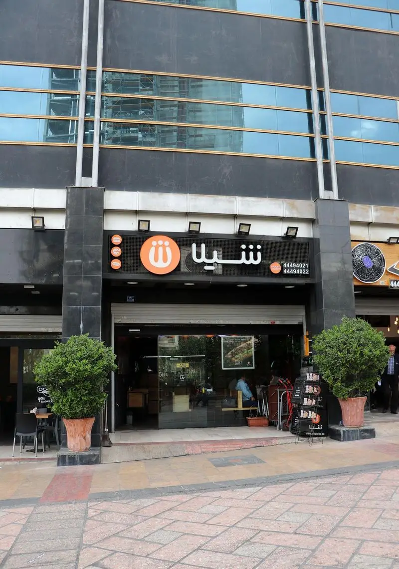 نمای اصلی و ورودی فست فود شیلا در یک ساختمان مدرن با شیشه‌های آبی بازتابنده. تابلوی بزرگ نارنجی شیلا، شماره‌های تماس ۴۴۴۹۴۰۲۱ و ۴۴۴۹۴۰۲۲ برای سفارش آنلاین.