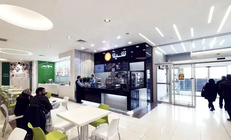 فضای داخلی مدرن فست فود شیلا با میز و صندلی‌های سبز و سفید، پیشخوان سیاه، کارمندان و مشتریان در حال خرید و صرف غذا