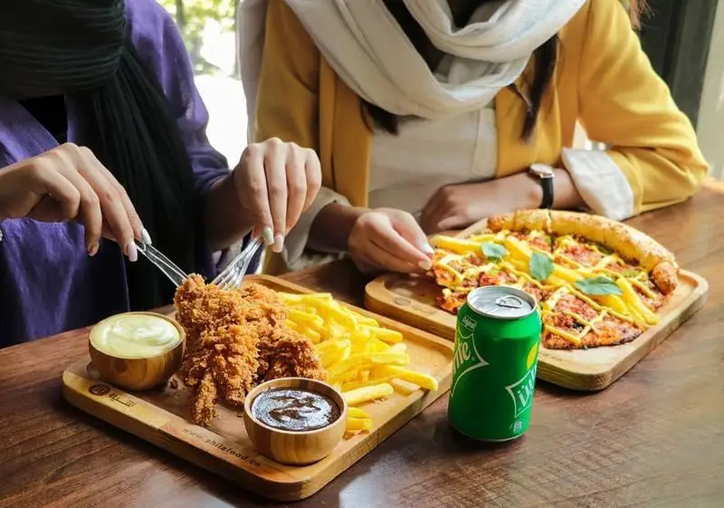 دو خانم در فست فود شیلا مشغول صرف غذای خوشمزه: مرغ سوخاری و سیب زمینی سرخ کرده با دو سس، در کنار پیتزای لذیذ با نوشابه.