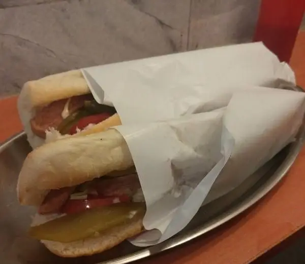 دو ساندویچ سوسیس و کالباس با نان باگت تازه، خیارشور و گوجه، همراه با سس، روی سینی فلزی از ساندویچی اغذیه فرد بلوار