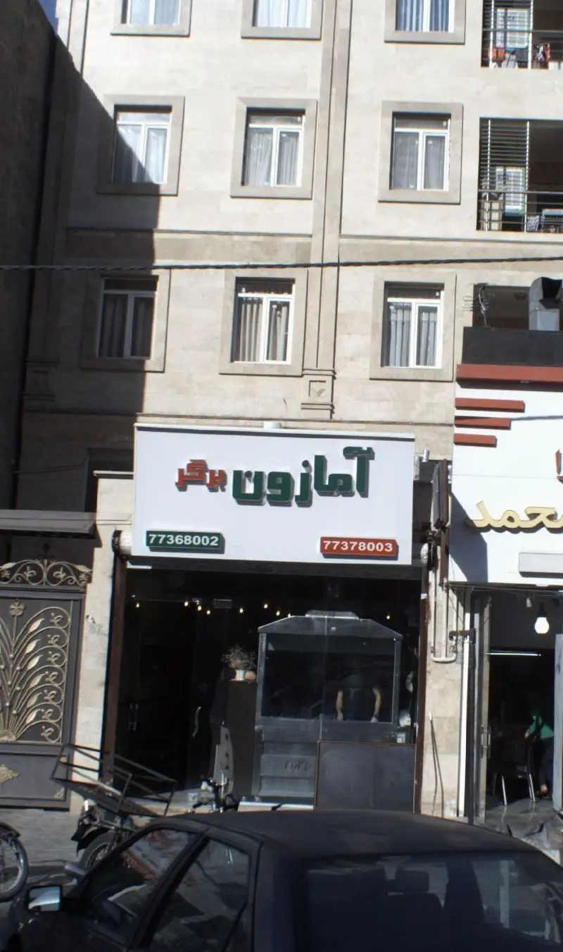 تصویری از نمای بیرونی فست فود امازون برگر در تهران، با تابلوی سبز و قرمز و شماره تلفن‌های 77368002 و 77378003.