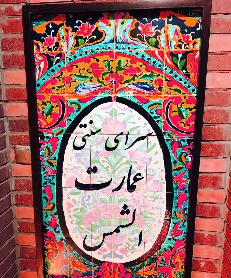 تصویری از تابلو کاشی‌کاری شده سنتی رستوران ایرانی عمارت شمس، با خط نستعلیق و طرح‌های گل و مرغ ایرانی، دیوار آجری.