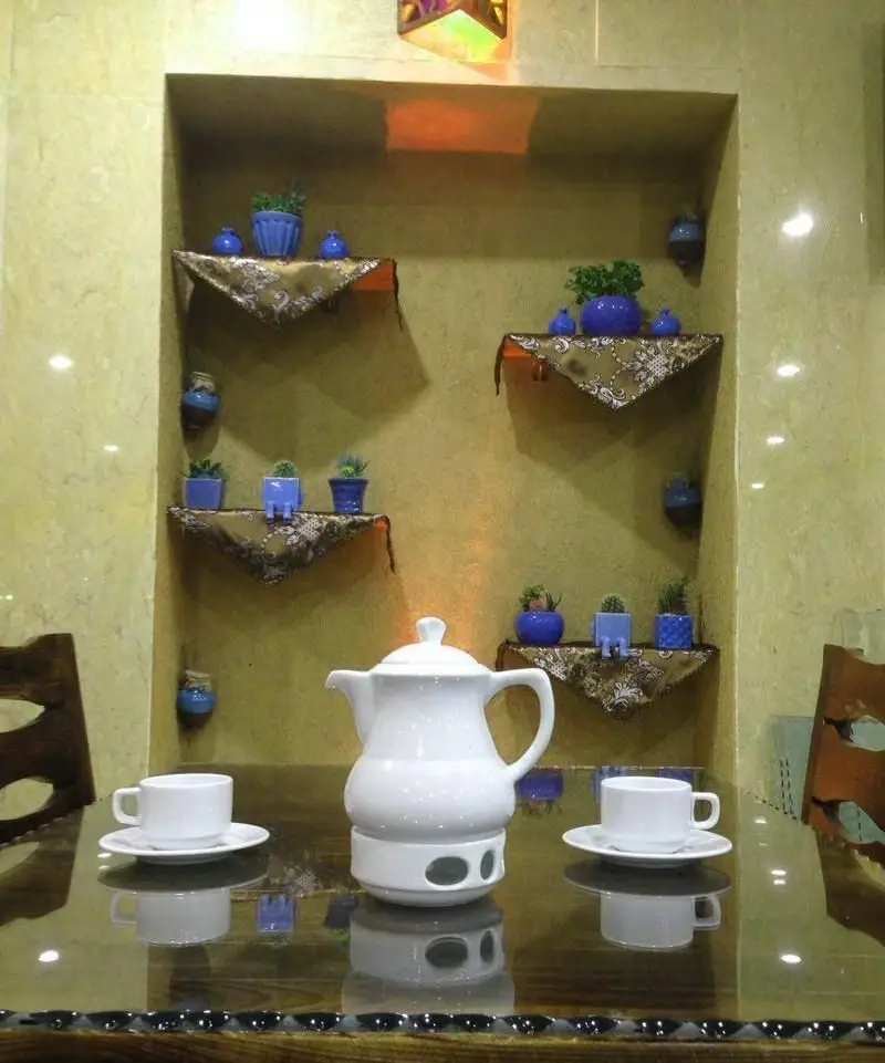 دکوراسیون سنتی و فضای دلنشین رستوران ایرانی عمارت شمس با چیدمان قوری و فنجان چای روی میز شیشه‌ای آماده پذیرایی.