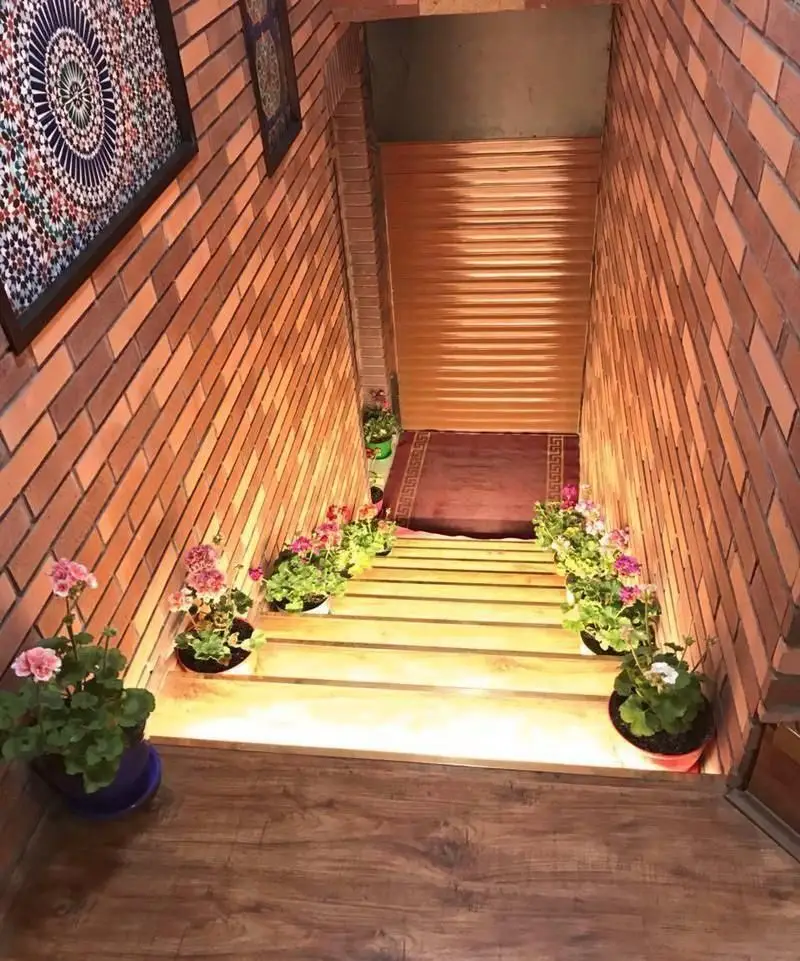 ورودی دنج و سنتی رستوران ایرانی عمارت شمس با دیوارهای آجری، پله‌های چوبی روشن، گلدان‌های گل و آثار هنری ایرانی، فضایی دلپذیر.