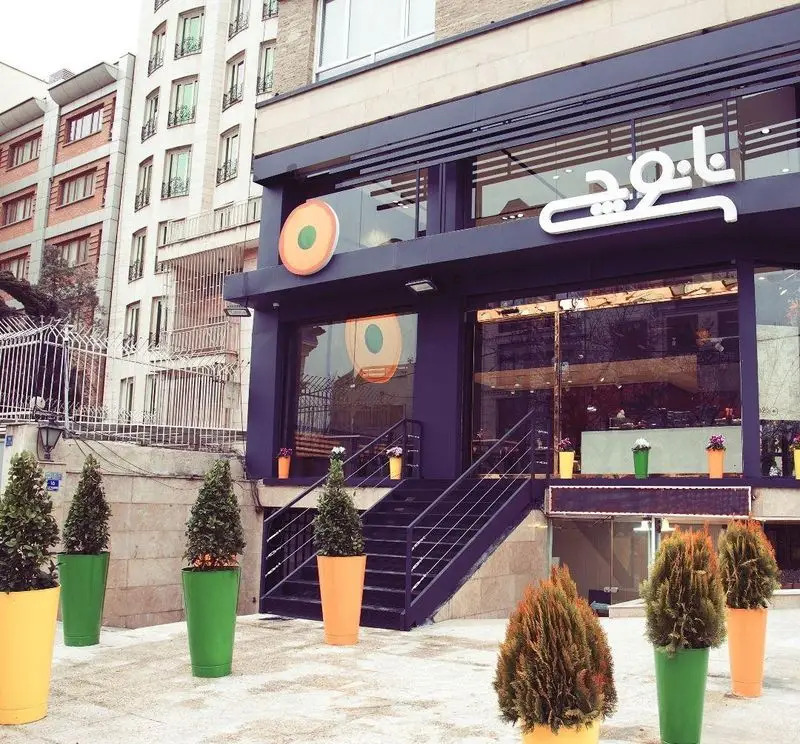 نمای بیرونی ساختمان رستوران نانوچی با ورودی پلکانی، گلدان‌های تزئینی رنگارنگ و تابلو نام برند، برای جذب مشتریان طراحی شده است.