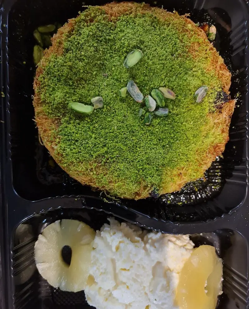 دسر کنافه پسته ای علی کنتیا با پسته سبز فراوان، سرو شده در ظرف با کرم و آناناس تازه. طعمی لذیذ و بی‌نظیر.