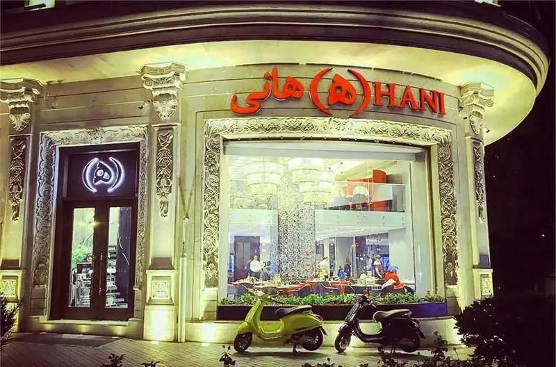 نمای شب رستوران ایرانی هانی، با معماری لوکس، نورپردازی دلنشین و دکوراسیون مدرن. اسکوترها در ورودی. لوگو هانی برجسته است.