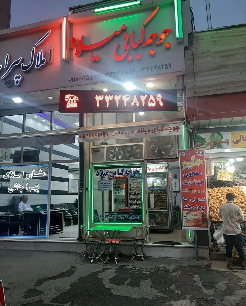 نمای بیرونی جوجه کبابی میلاد، رستوران سرو کننده کباب با تابلوهای نئونی درخشان و صندلی‌های بیرون، در کنار بنگاه املاک و فروشگاه میوه.