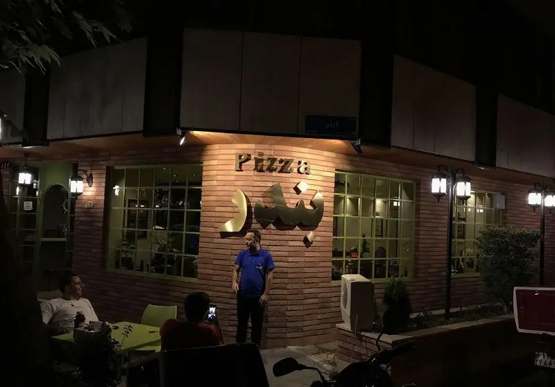 نمای بیرونی پیتزا بندر در شب با نورپردازی گرم و مشتریان نشسته، یک مکان دنج و عالی برای تجربه طعم پیتزا در فضایی دلنشین.