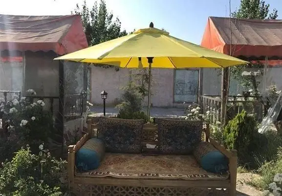 نمایی از فضای باز سنتی رستوران چهلستون، شامل تخت چوبی با قالیچه و کوسن زیر چتر زرد، احاطه شده با فضای سبز و گل.