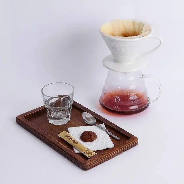 صحنه‌ای از آماده‌سازی قهوه دمی تخصصی در کافه و کافی شاپ لیدو پلاس، شامل ابزار دم‌آوری قهوه، سینی پذیرایی چوبی با لیوان آب، قاشق، شکر و کوکی شکلاتی.