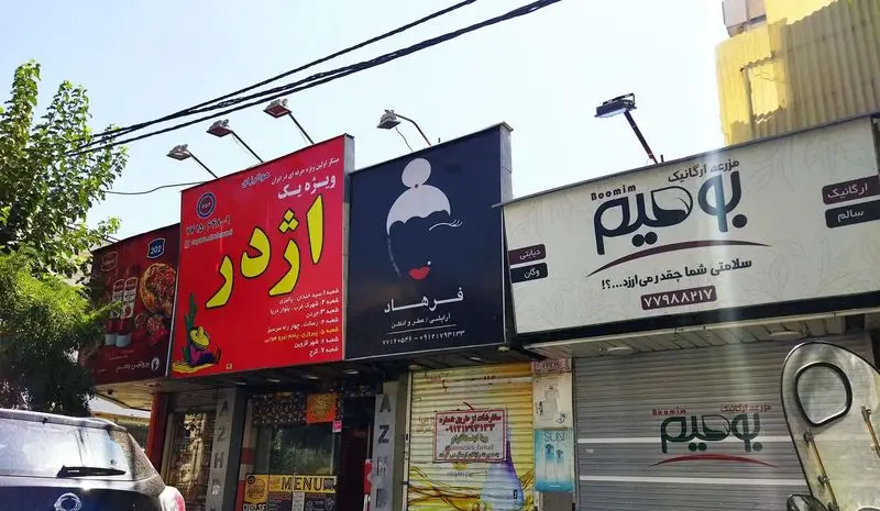 تابلوی پرنور فست فود اژدر زاپاتا (ویژه یک)، با زمینه قرمز و نوشته زرد، همراه با لیست شعب در کنار مغازه های دیگر.