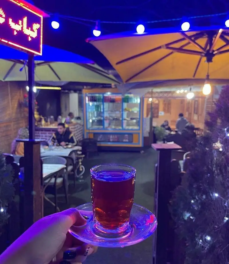 چای داغ در دست مشتری در شب زیبای جگرسرای کلبه صحرا، تجربه‌ای دلنشین در فضای باز و سنتی، مکانی ایده‌آل برای کباب.