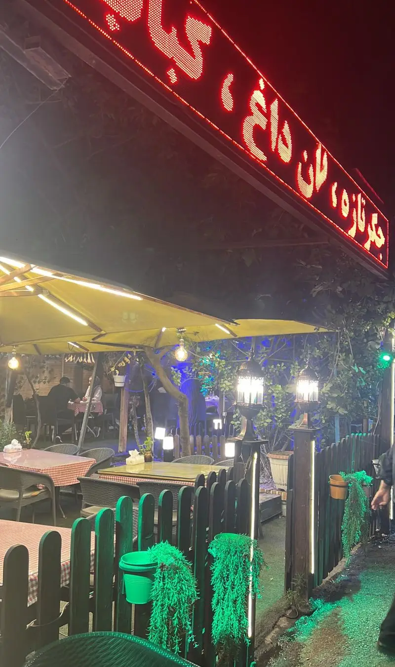 نمایی شبانه از فضای باز جگرسرای کلبه صحرا با چیدمان دلنشین میز و صندلی، سایبان و نورپردازی گرم برای تجربه غذای ایرانی.