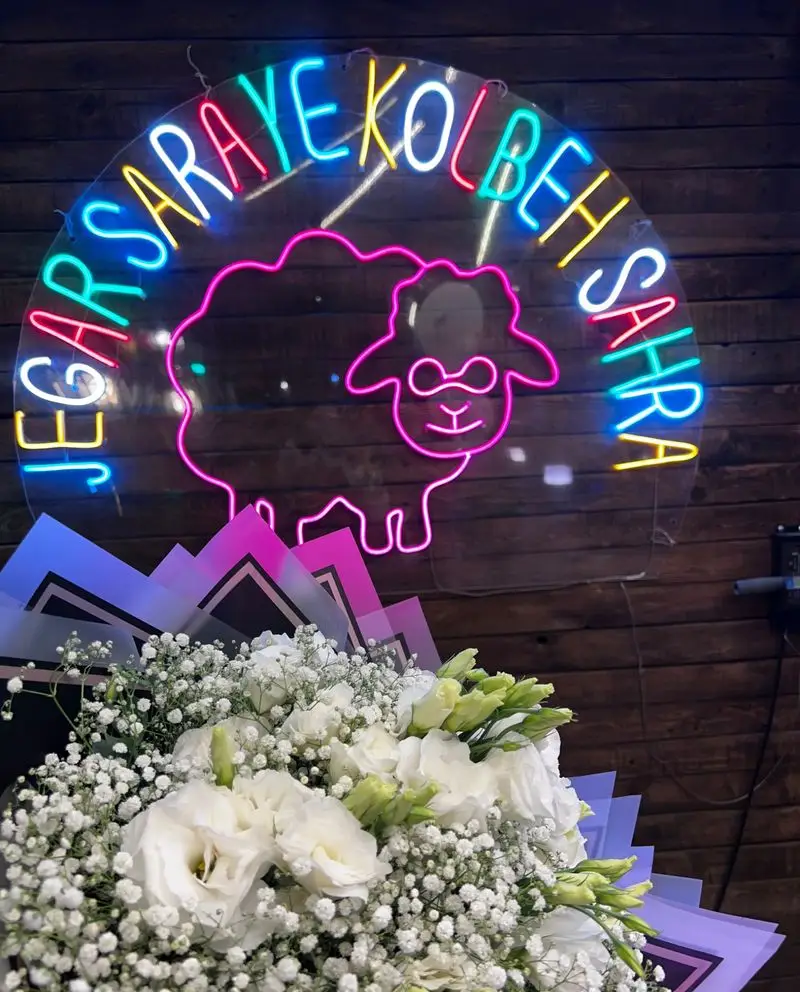 تابلو نئونی جذاب جگرسرای کلبه صحرا با نوشته‌های رنگی و طرح گوسفند، روی دیوار چوبی، همراه با دسته‌گل سفید شیک در جلو.