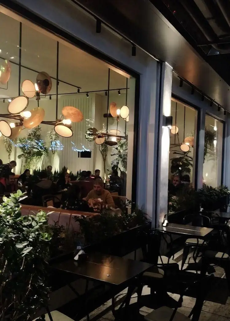 نمای شبانه رستوران جو با دکوراسیون داخلی شیک، نورپردازی مدرن، گیاهان سبز و مشتریانی که در حال صرف شام در فضایی دلنشین هستند.