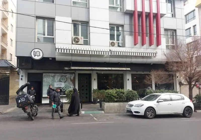 نمای خارجی رستوران جو با طراحی مدرن، تابلوی "جو" و لوگوی سفید، پیاده‌رویی با رهگذران و موتور سیکلت دلیوری، خودروی پارک شده در خیابان.