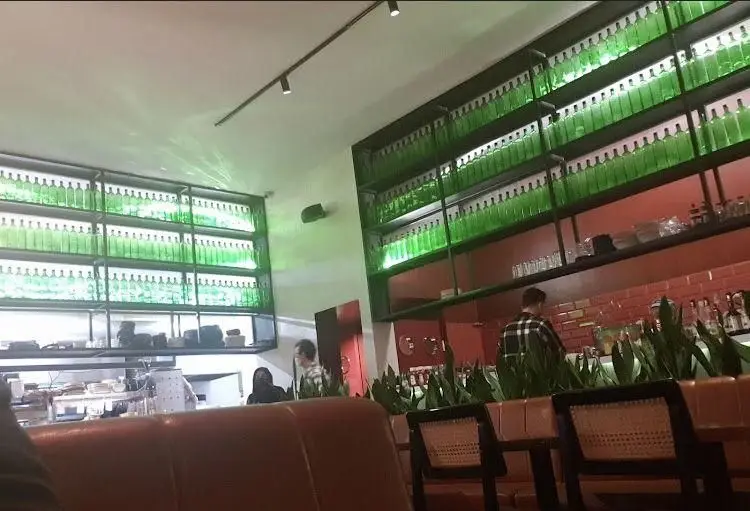 فضای داخلی رستوران جو با قفسه‌های بلند پر از بطری‌های سبز نورانی، دیوارهای آجری قرمز و گیاهان زینتی. دکوراسیون منحصر به فرد.