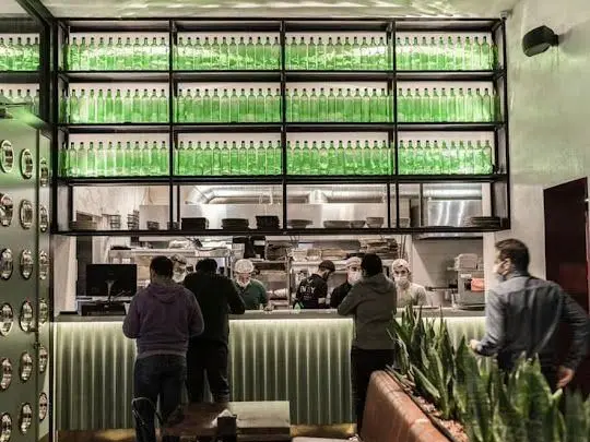 فضای داخلی رستوران جو با دکوراسیون مدرن، آشپزخانه باز و قفسه‌های بطری‌های سبز. محیطی پویا با حضور مشتریان و سرآشپزهای مشغول به کار.