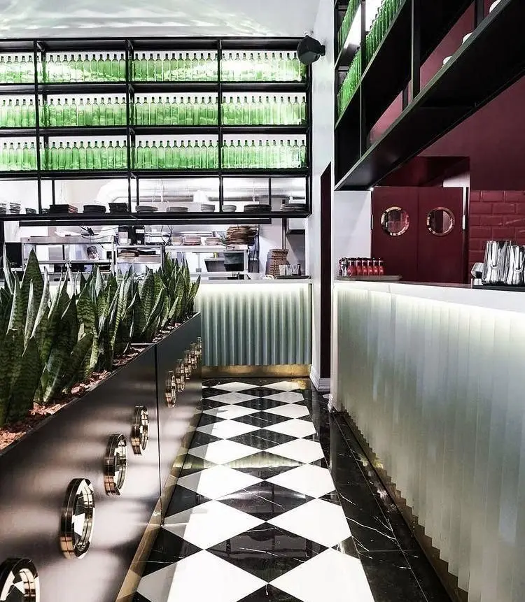 دکوراسیون شیک و مدرن رستوران جو شامل قفسه‌های دیواری فلزی با بطری‌های سبز، بار نوشیدنی نورانی، کفپوش شطرنجی و گلدان‌های گیاه سانسوریا.