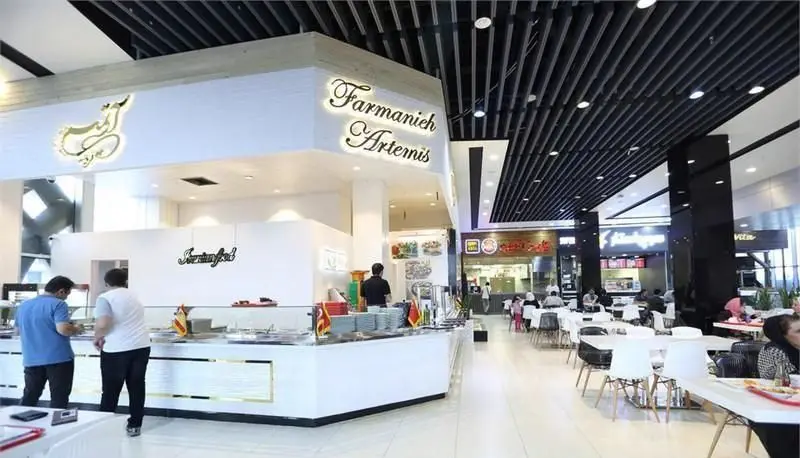تصویری از فضای داخلی فوکورت آرتمیس با پیشخوان بزرگ Farmanieh Artemis، مشتریانی در حال سفارش و میز و صندلی‌های چیده شده در یک محیط مدرن و روشن.