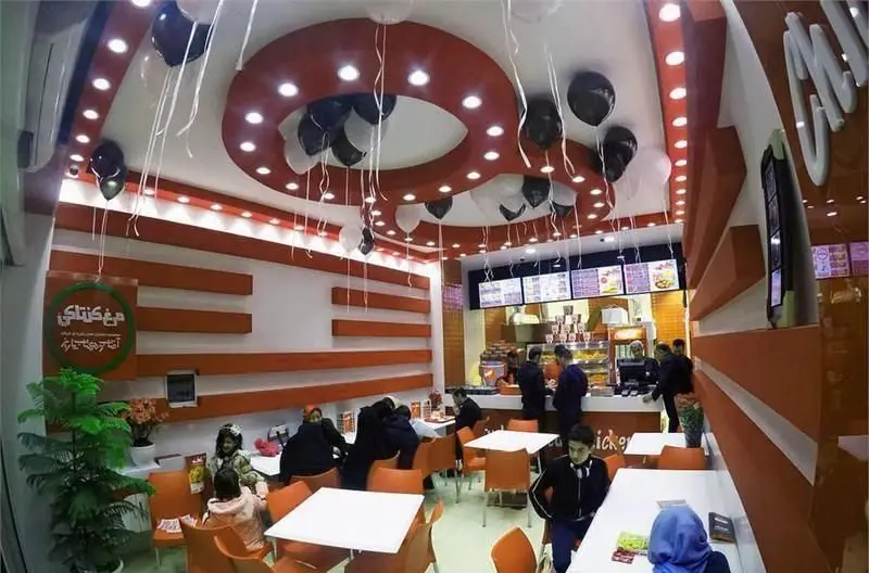 فضای داخلی شعبه کی اف سی هروی با دکوراسیون مدرن نارنجی و سقف تزئین شده با بالون، مشتریان در حال سفارش و صرف غذا.