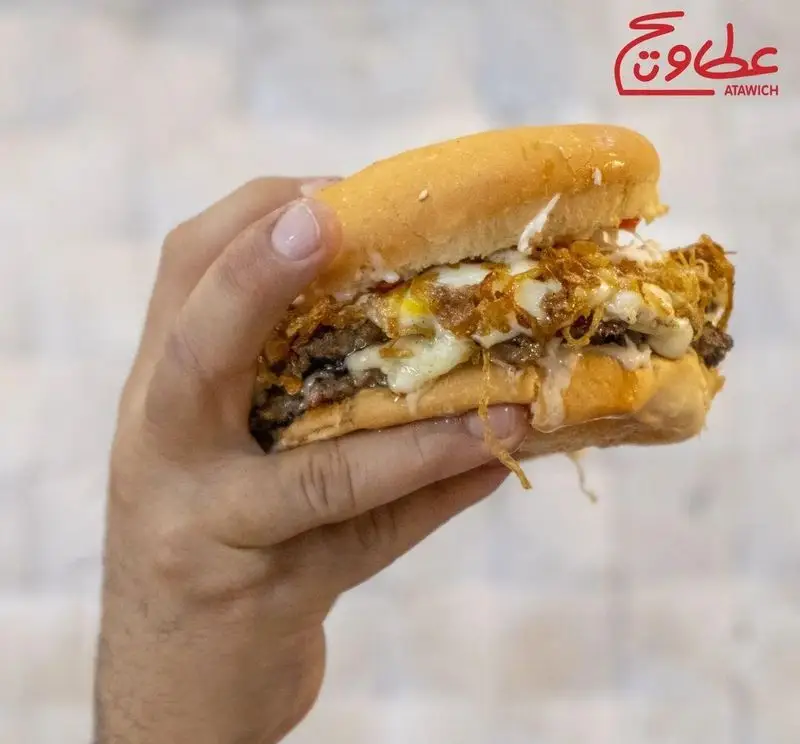 ساندویچ برگر ویژه عطاویچ فردوس غرب با دو لایه گوشت لذیذ، پنیر ذوب شده، تخم مرغ و پیاز داغ ترد، در دست مشتری.