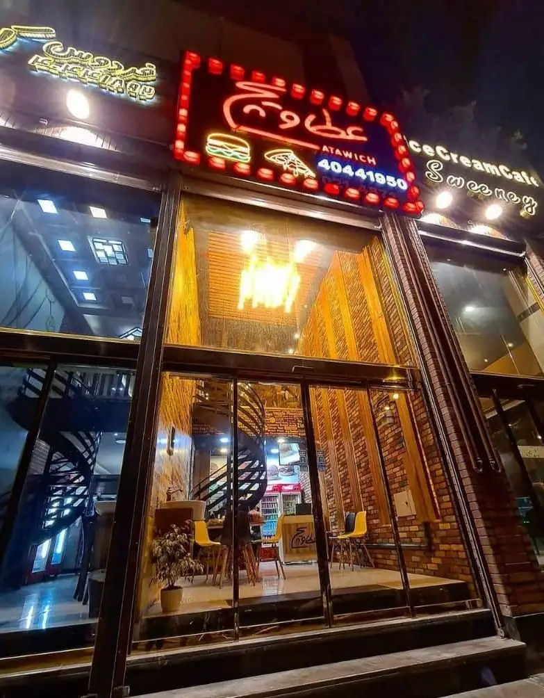 رستوران عطاویچ فردوس غرب: نمای شبانه ساختمان با ورودی شیشه‌ای، تابلوهای نئون روشن برای همبرگر، پیتزا و شماره تماس. پله مارپیچ داخلی.