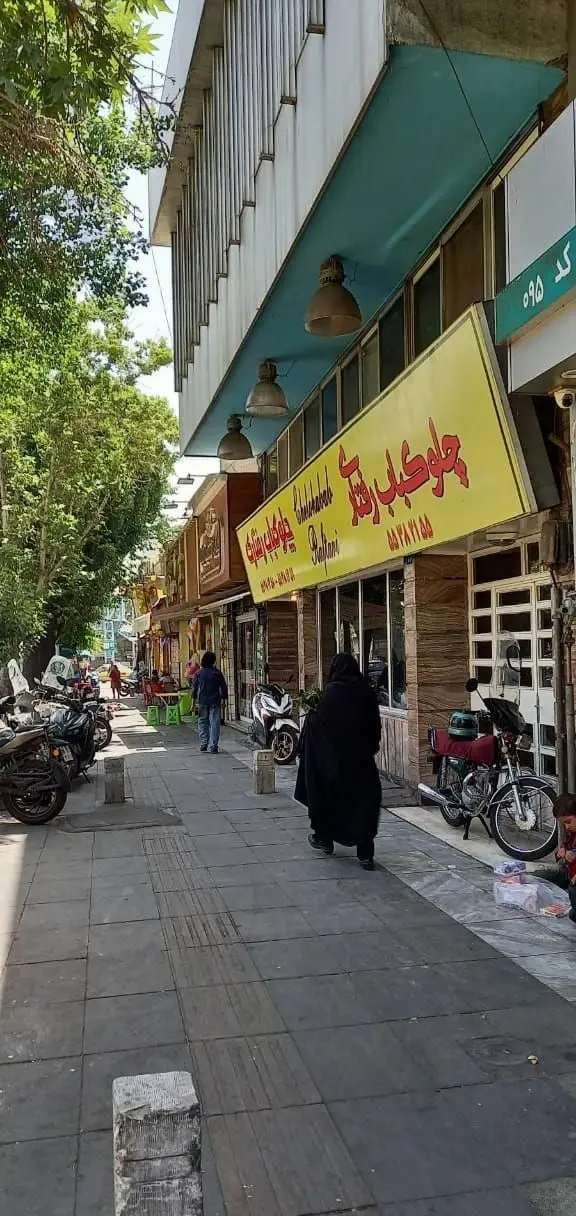 نمای بیرونی رستوران چلوکباب رفتاری، با تابلوی زرد و پرنور، عابران پیاده و موتورسیکلت‌ها در یک خیابان شلوغ تهران.