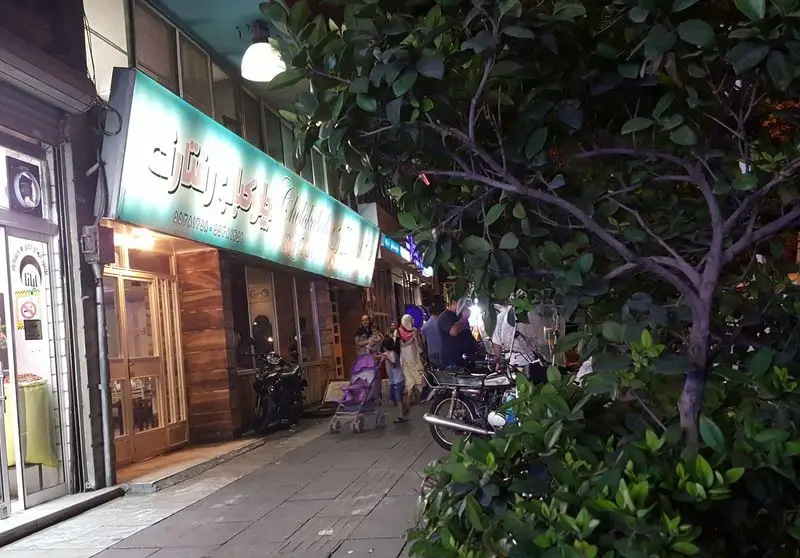 نمای شبانه رستوران رفتاری با تابلوی روشن و مشتریان در پیاده‌رو. فضایی دلپذیر برای تجربه طعم لذیذ غذاهای ایرانی.