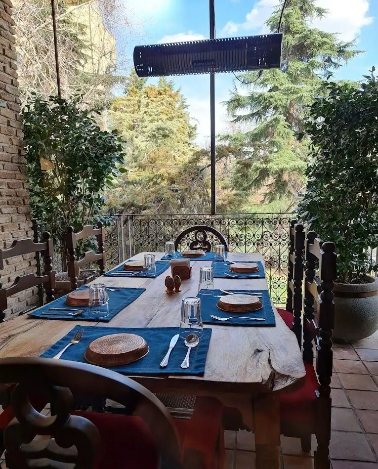 میز چوبی بزرگ با چیدمان زیبا در تراس رستوران مس توران، شامل بشقاب مسی و دستمال آبی، با منظره درختان و آسمان آبی