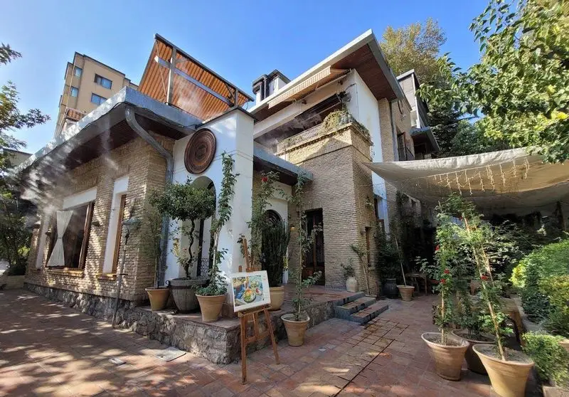 نمای بیرونی رستوران مس توران با معماری سنتی و مدرن، حیاط سرسبز، گلدان‌ها، آب‌پاش‌ها و فضای دنج برای تجربه غذایی دلنشین.