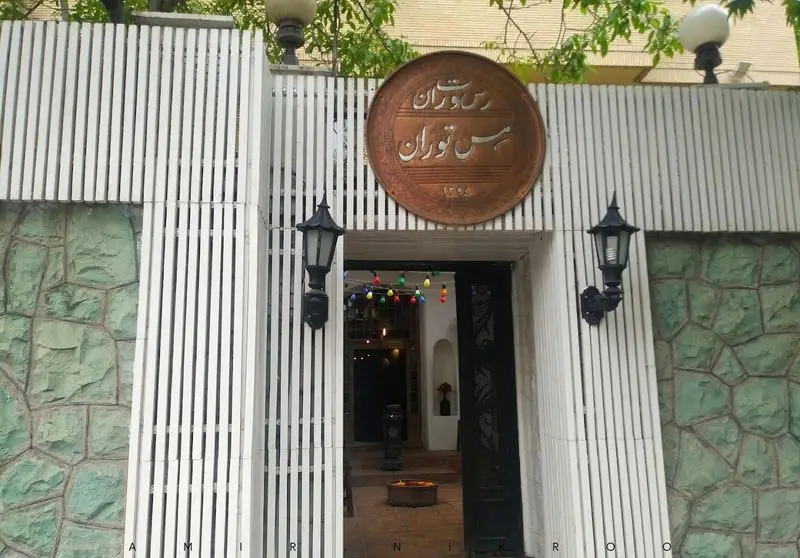 ورودی جذاب و نمای بیرونی رستوران مس توران، با تابلوی مسی اصیل، نرده‌های سفید و دیوارهای سنگی سبز، مکانی خاص برای ناهار و شام.