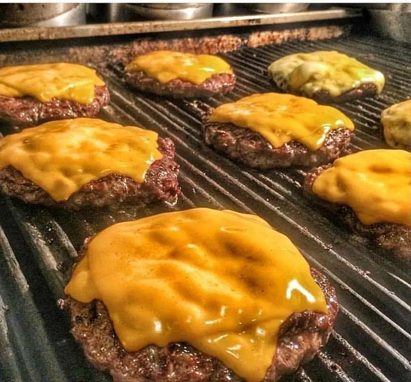 همبرگرهای پنیری فست فود اژدر زاپاتا در حال گریل شدن روی توری داغ، با پنیر چدار آب شده و طلایی، تجربه‌ای لذیذ.