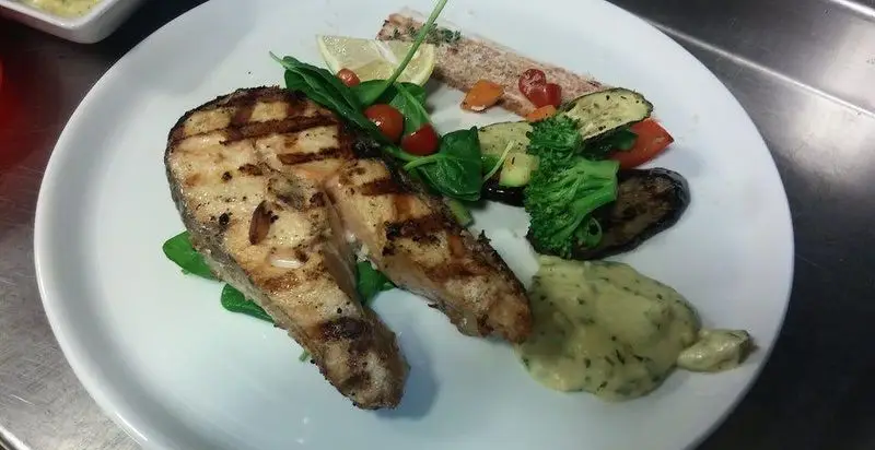 یک بشقاب غذای لذیذ شامل استیک ماهی گریل شده، همراه با سبزیجات تازه، اسفناج، گوجه گیلاسی و سس مخصوص رستوران فرسکا.